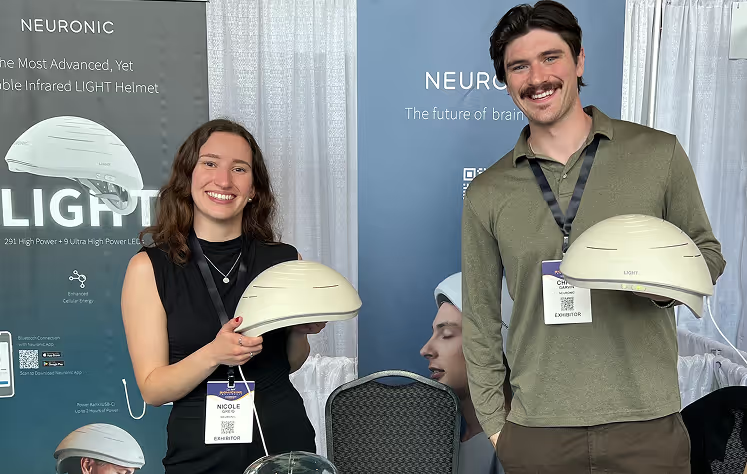Two people holding Neuronic Helmets at a product display booth, both smiling at the camera