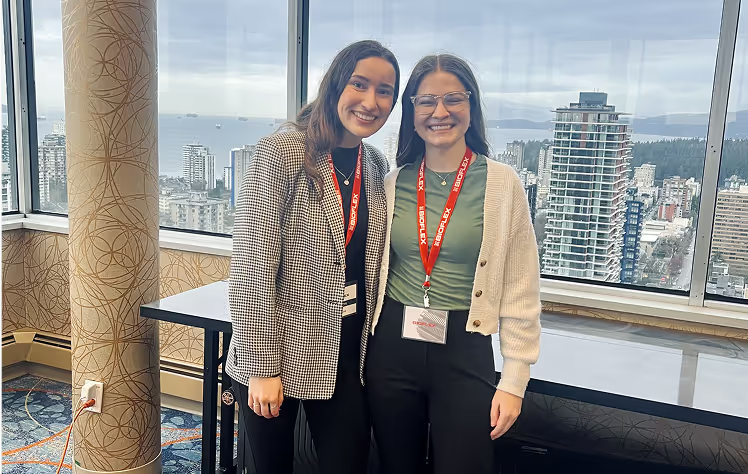 Two women smiling at camera in high-rise office with city skyline view, both wearing red lanyards with badges