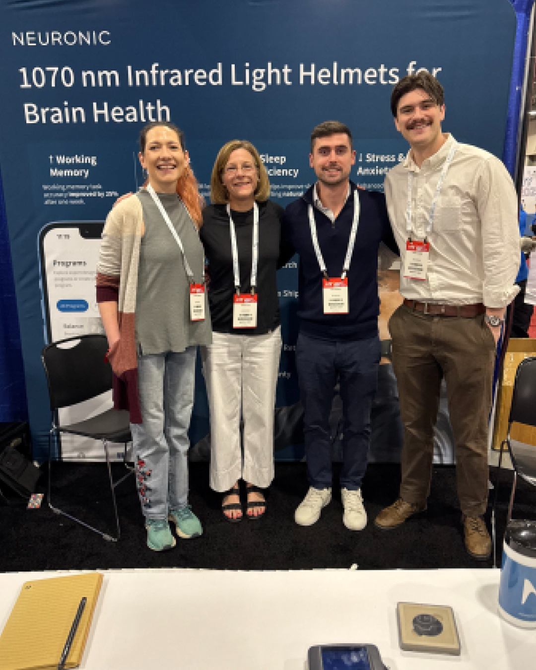 Four people wearing conference badges standing in front of a Neuronic booth displaying "1070 nm Infrared Light Helmets for Brain Health