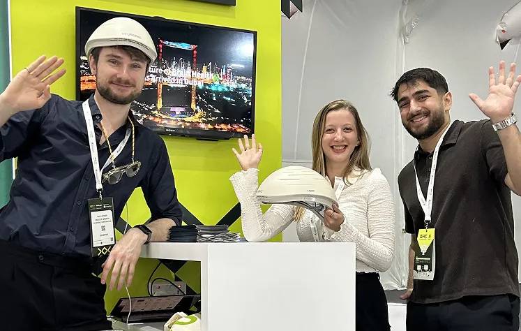 Three people at a trade show booth waving at the camera, woman in center holding the Neuronic Helmet