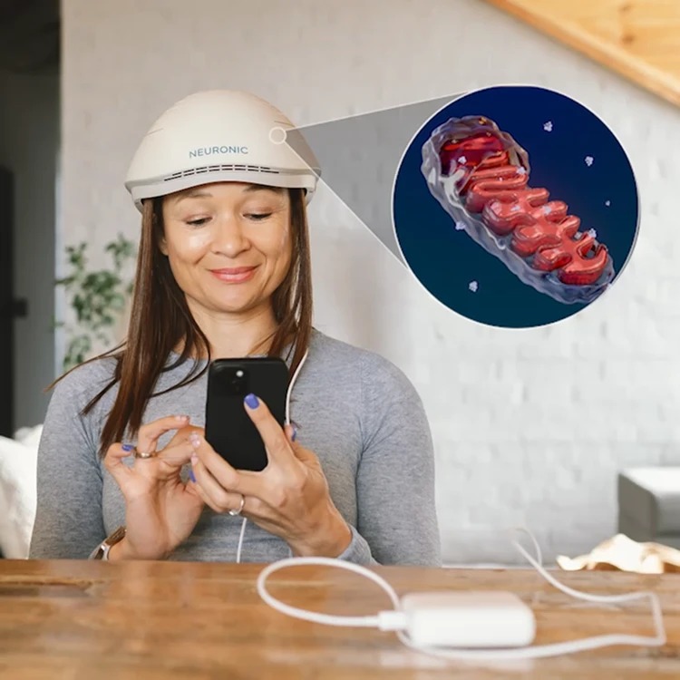 A woman wearing the Neuronic LIGHT at home while using her phone, with a mitochondrion shown enlarged beside her.