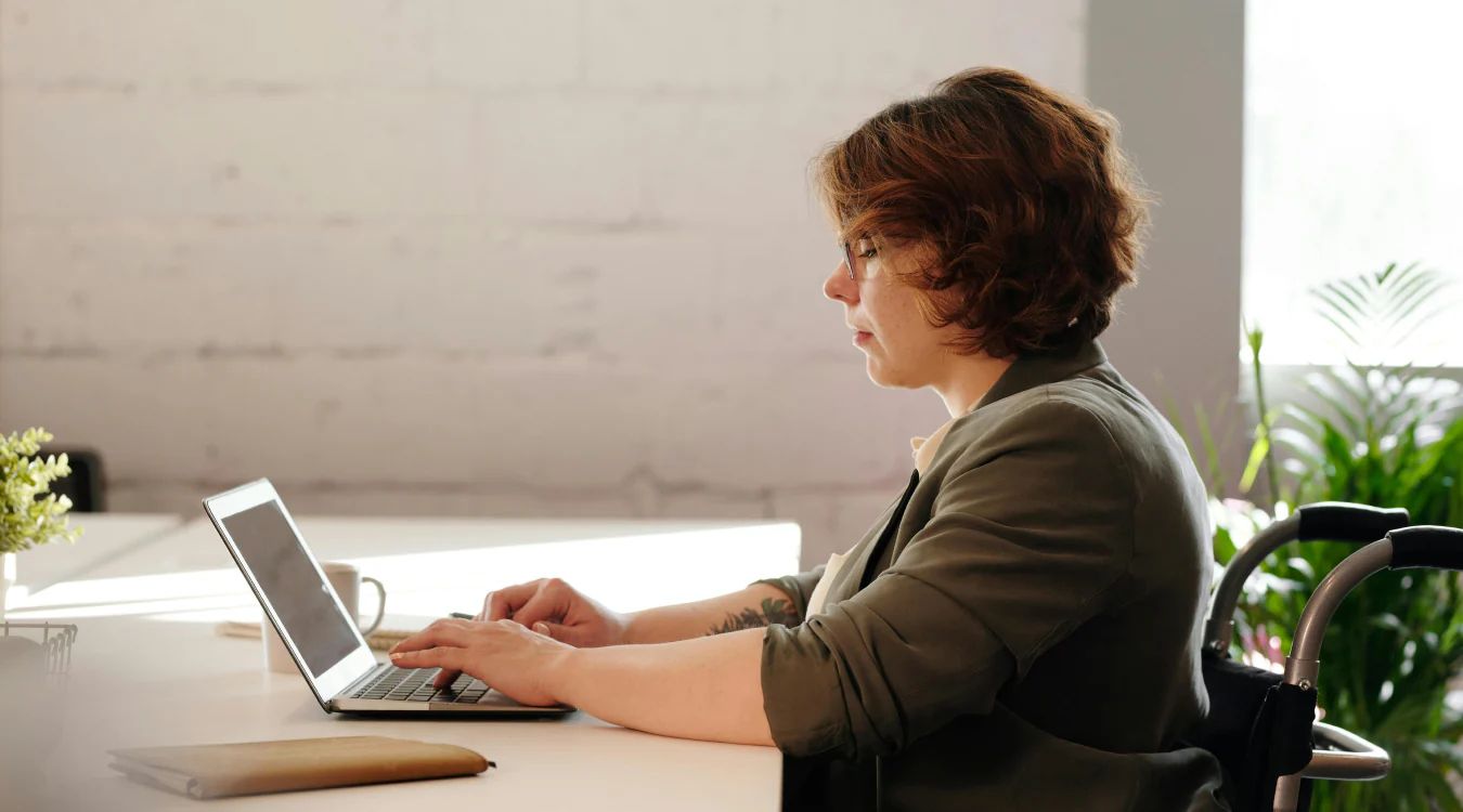 Person with reddish-brown hair wearing olive jacket, seated at desk with laptop and coffee cup, looking to the side