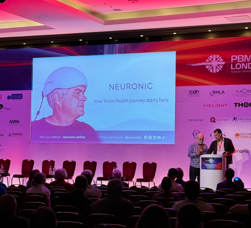 Man wearing Neuronic Helmet on conference stage screen, with text "Your brain health journey starts here" and two presenters at podium below