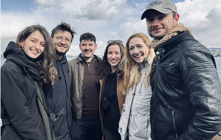 Group of six people smiling outdoors against a cloudy sky