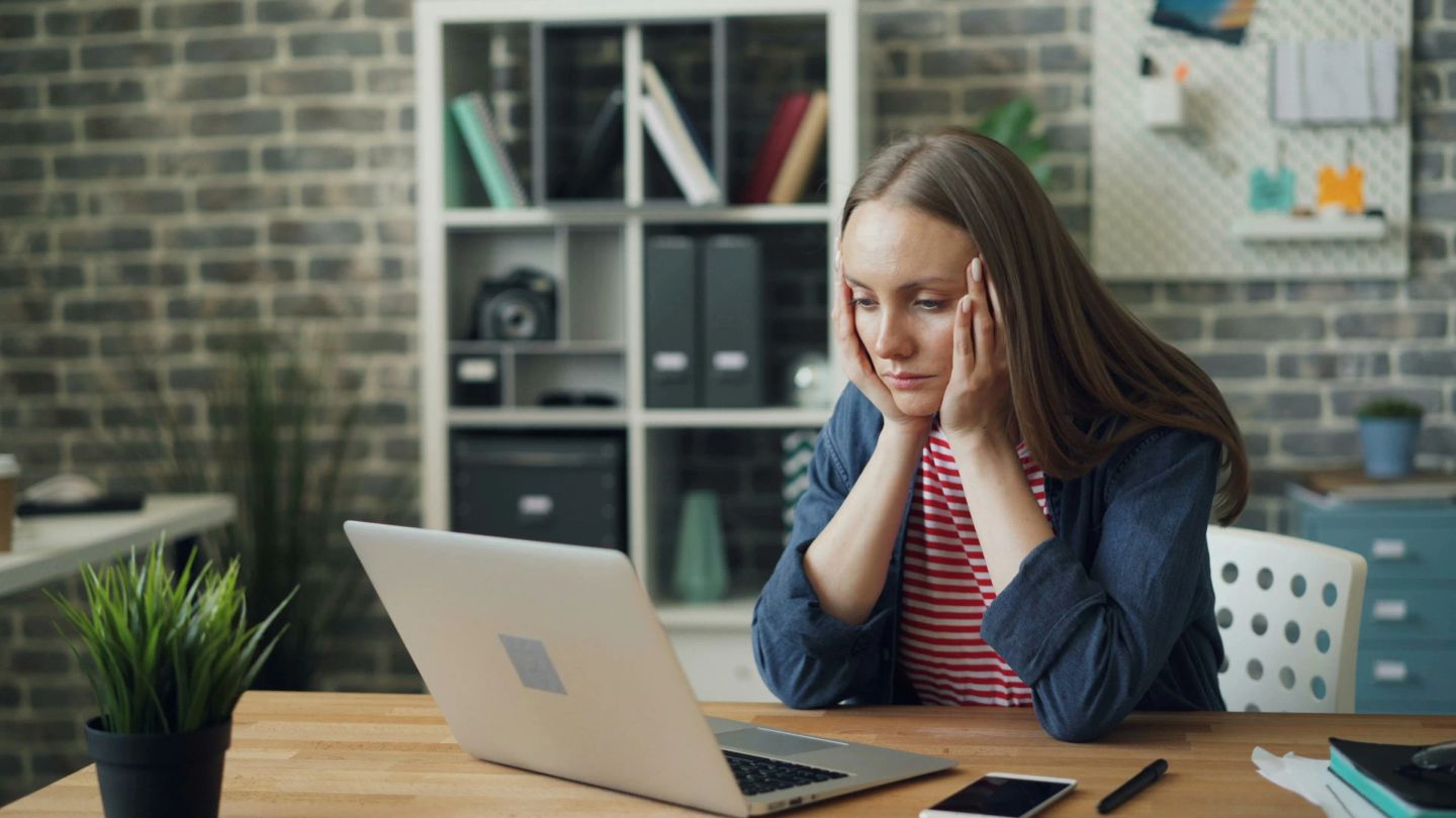 Woman at desk looking at laptop screen with hands to her head, appearing tired or stressed