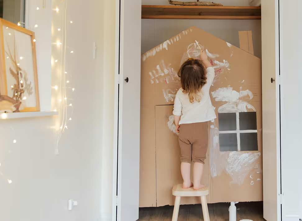 Young child standing on wooden stool, drawing on large cardboard house structure with white chalk