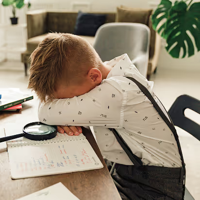 Child leaning over desk with magnifying glass, examining notebook, with pencils and sketches visible