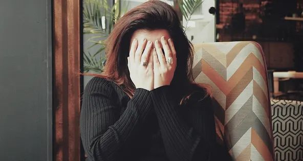 Person with hands covering their face, sitting on a patterned chair wearing a dark pinstriped shirt