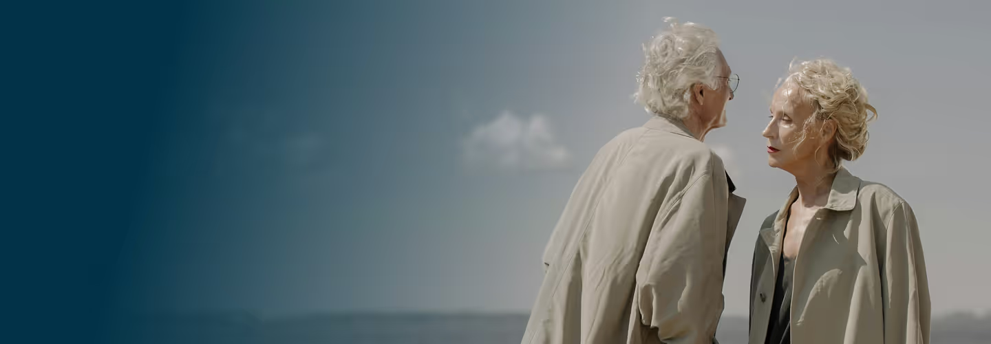Two older adults standing outdoors against a cloudy sky, wearing light-colored jackets