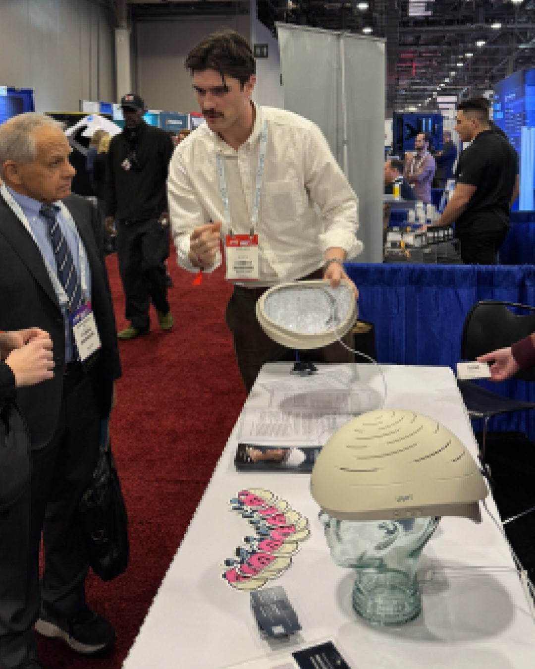 Man in white shirt demonstrating Neuronic Helmet at trade show booth, holding device above table display with other light-therapy equipment