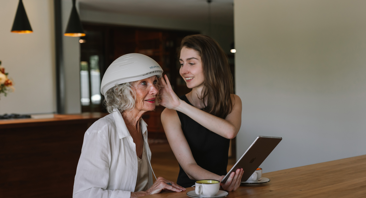 Older woman wearing Neuronic Helmet, seated at table with younger woman holding tablet, both smiling