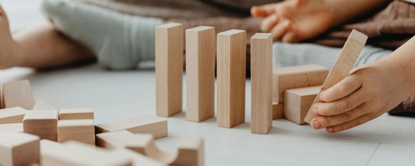 Child's hand placing wooden block next to standing blocks on white surface, adults in background