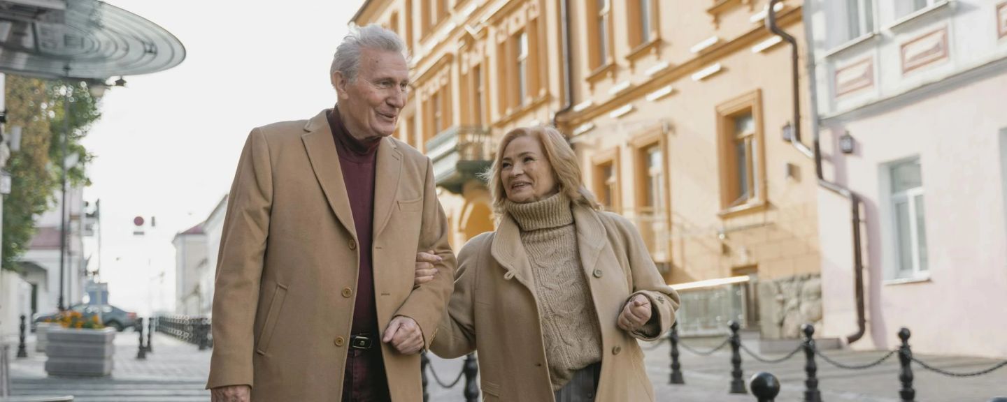 Two older adults walking arm-in-arm down a European city street lined with historic buildings