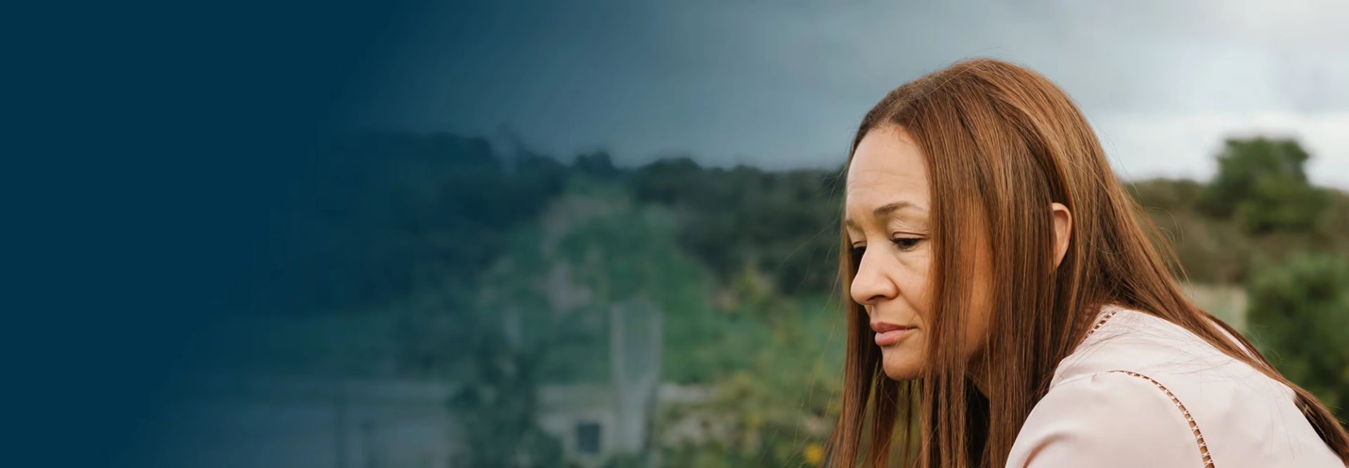 Woman with reddish-brown hair looking downward, with a blurred landscape of trees and hills in the background