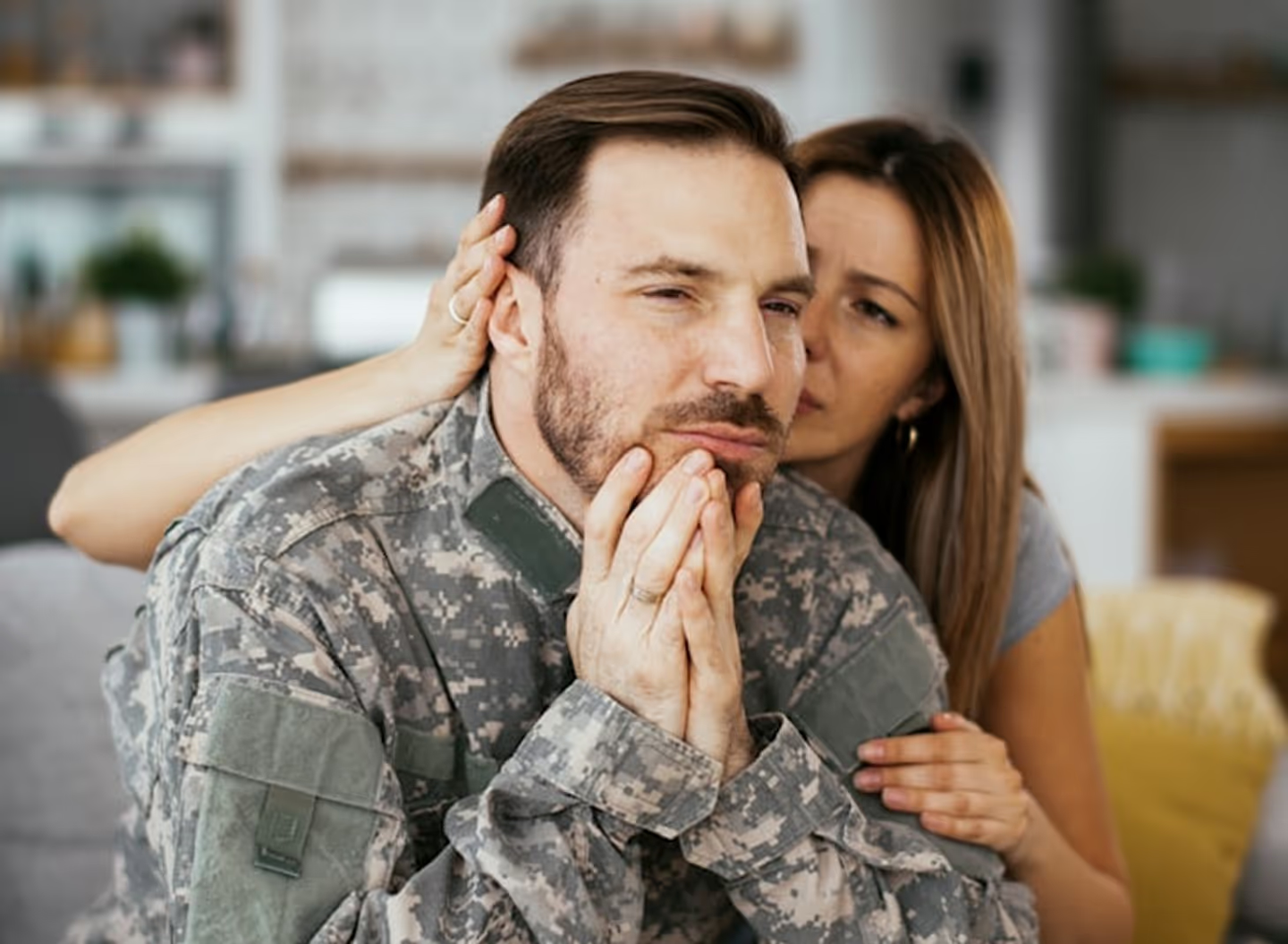 Man in military uniform looking contemplative while woman embraces him from behind in a kitchen