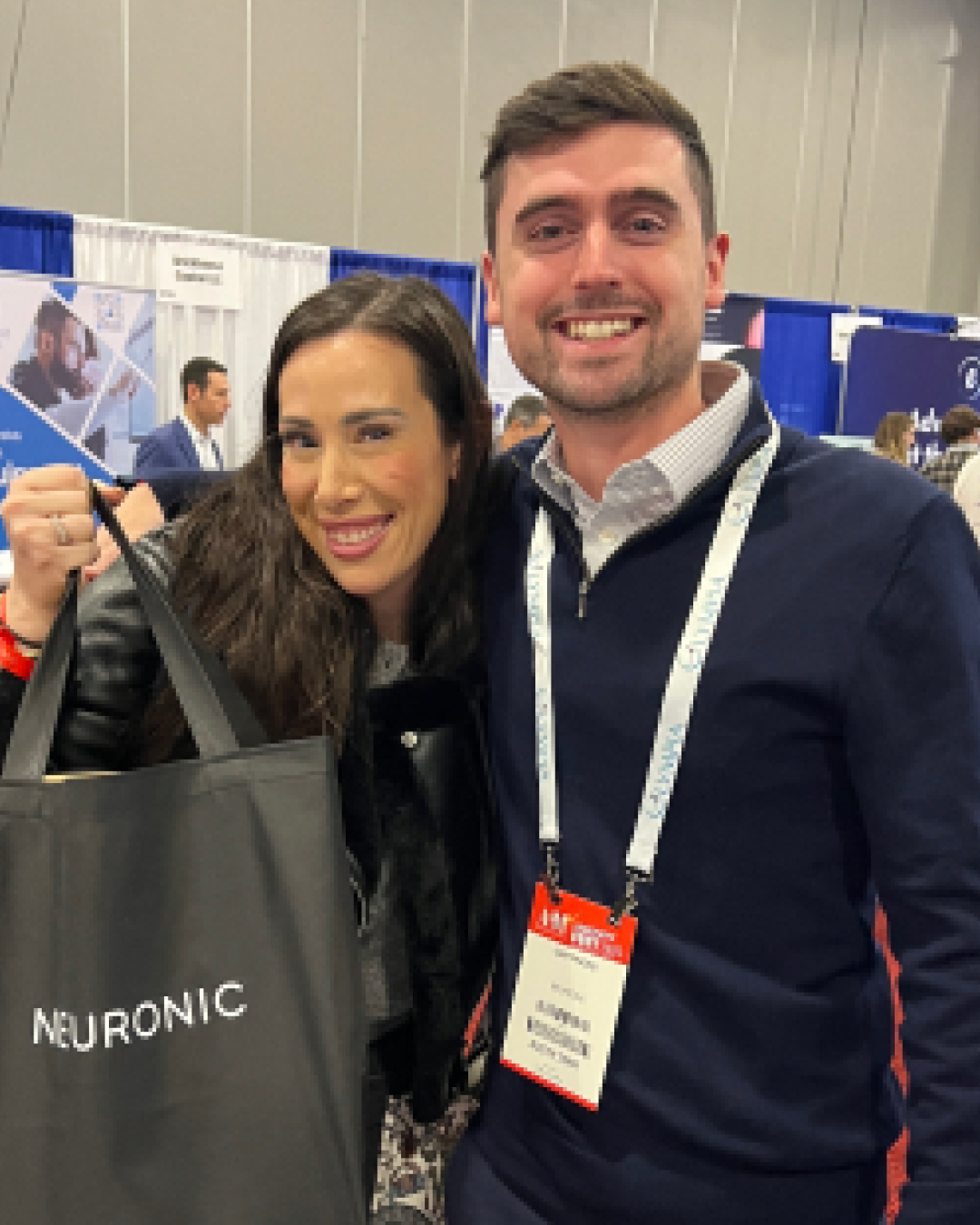 Two people smiling at a conference booth with Neuronic branding visible on a display behind them