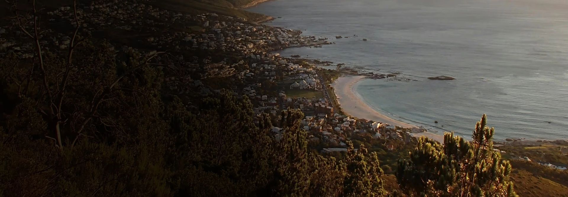 Aerial view of a coastal town nestled between forested hills and a curved sandy beach bordered by calm water