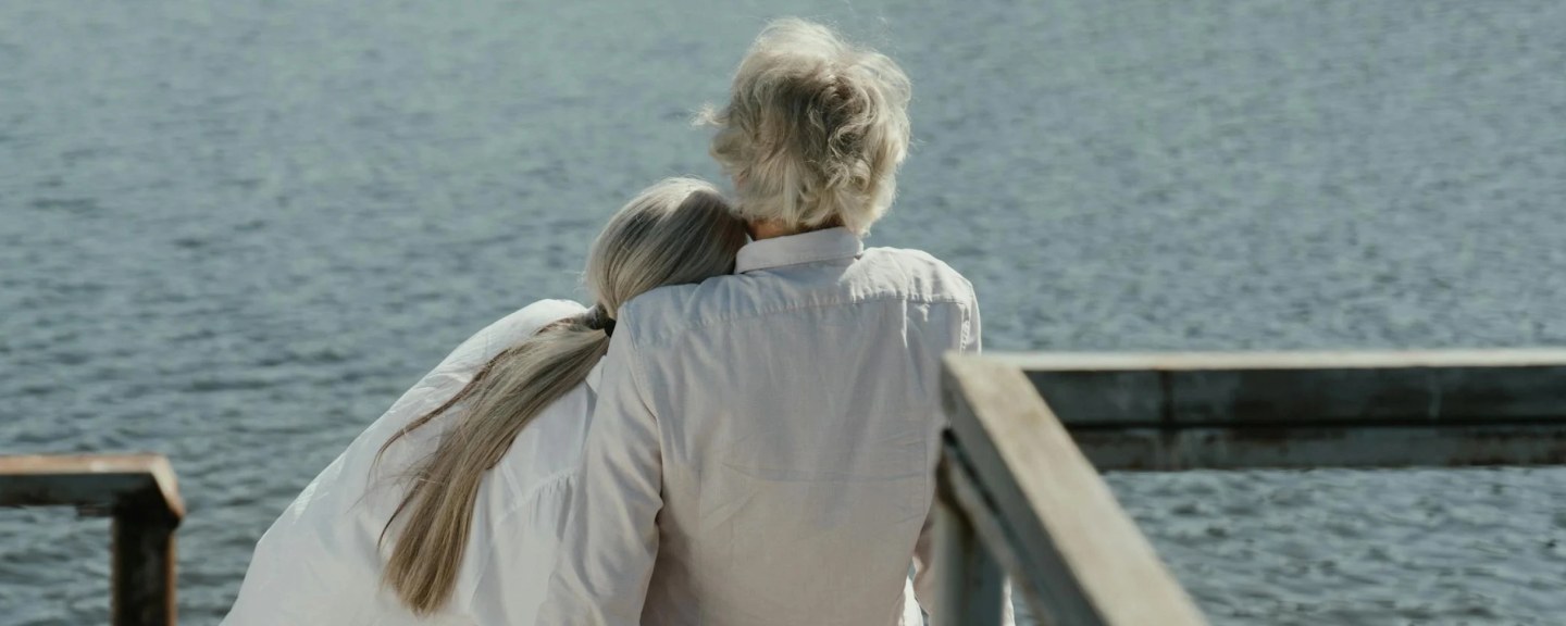 Older person with gray hair and younger person with long brown hair embracing while looking out at water from a pier
