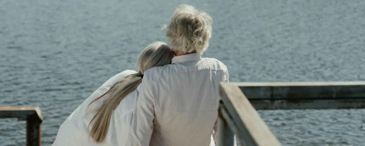 Older person with gray hair and younger person with long brown hair embracing while looking out at water from a pier