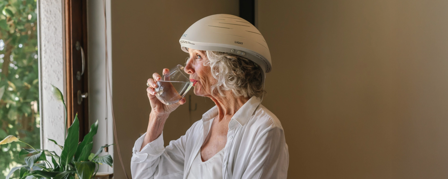 Older woman wearing Neuronic Helmet, drinking water from a glass