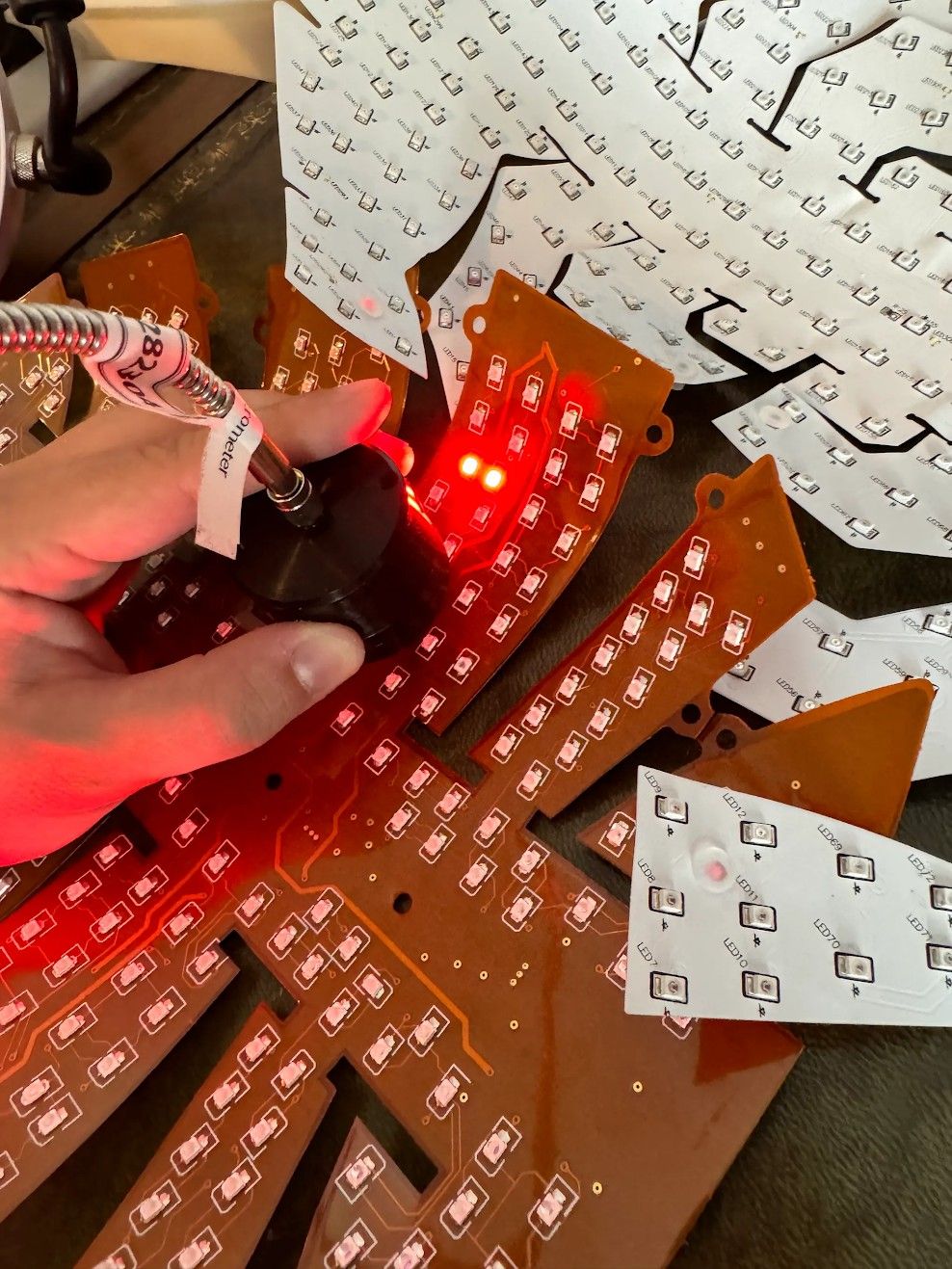 Hand holding connector cable above red LED circuit boards with illuminated lights, schematic diagrams visible in background