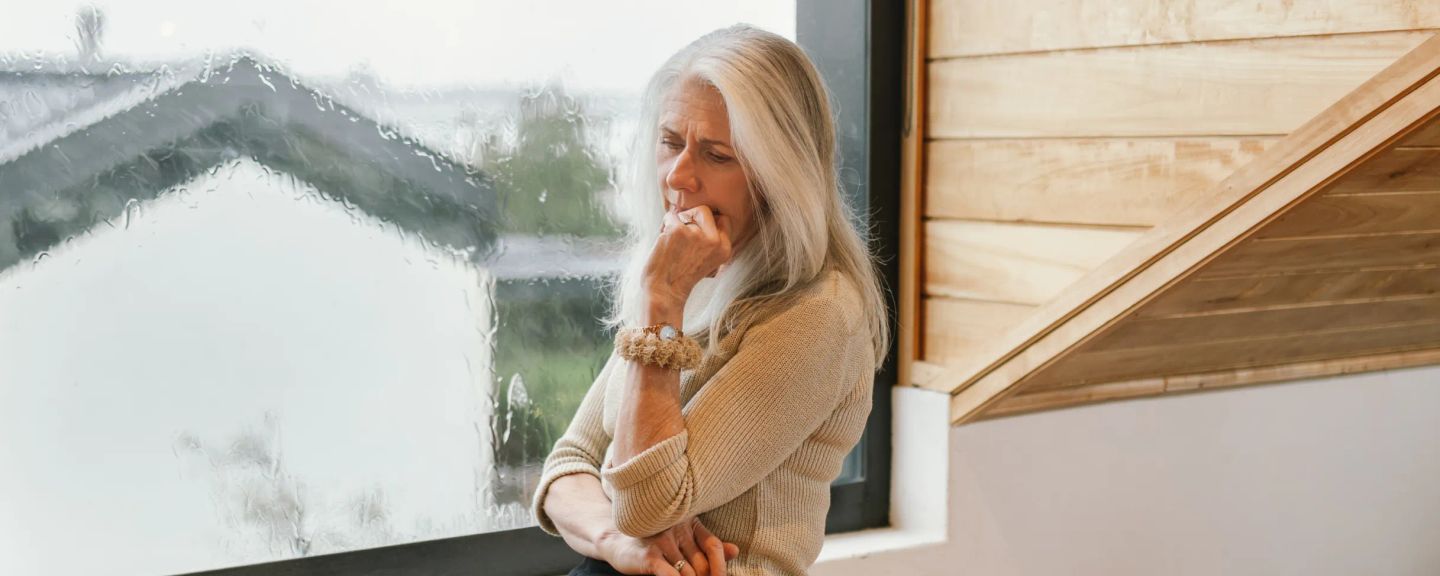 Woman with white hair wearing a beige sweater, hand to face, looking out a window at a snowy mountain landscape