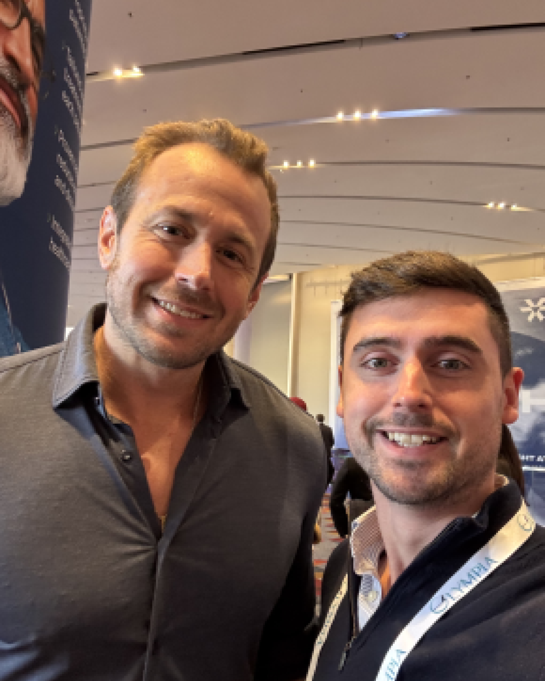 Two men smiling at the camera indoors at what appears to be a conference or event, with recessed ceiling lights visible in the background