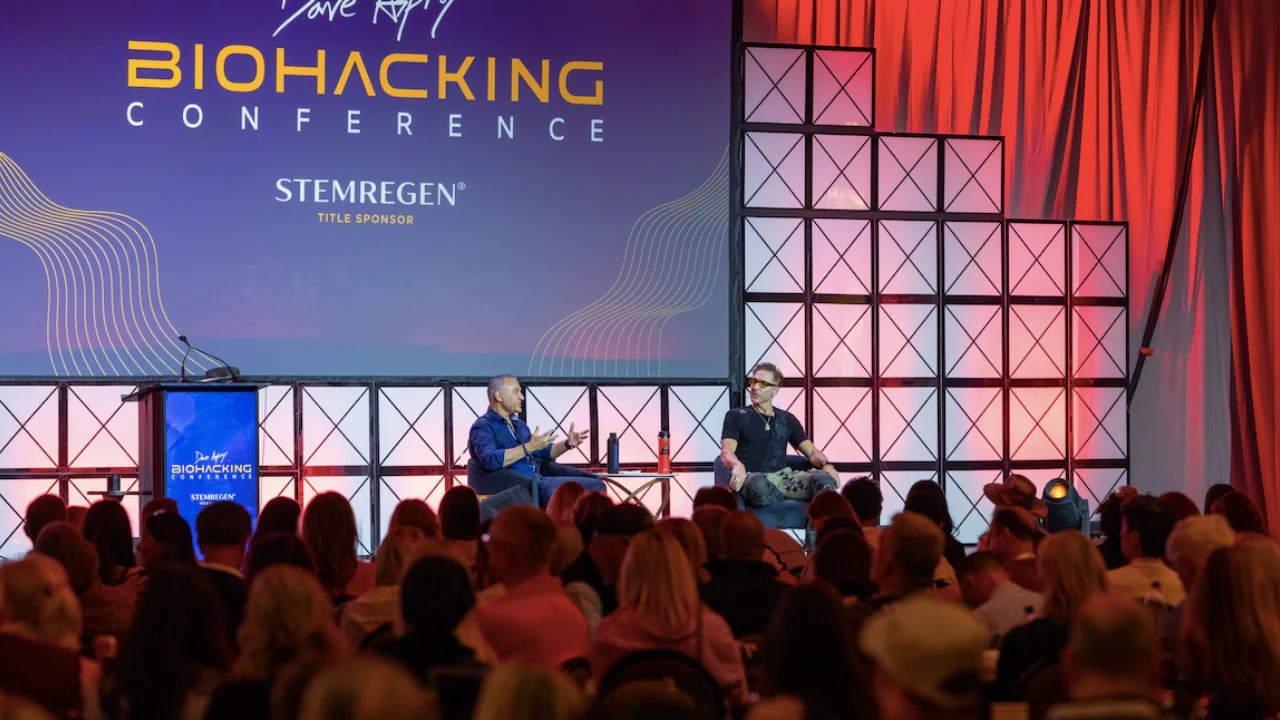 Two speakers on stage during Dave Asprey Biohacking Conference, audience in foreground, purple and red lighting, Stemregen title sponsor displayed