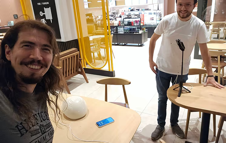 Man with long dark hair wearing Neuronic Helmet, smiling at table in modern showroom while another man stands nearby
