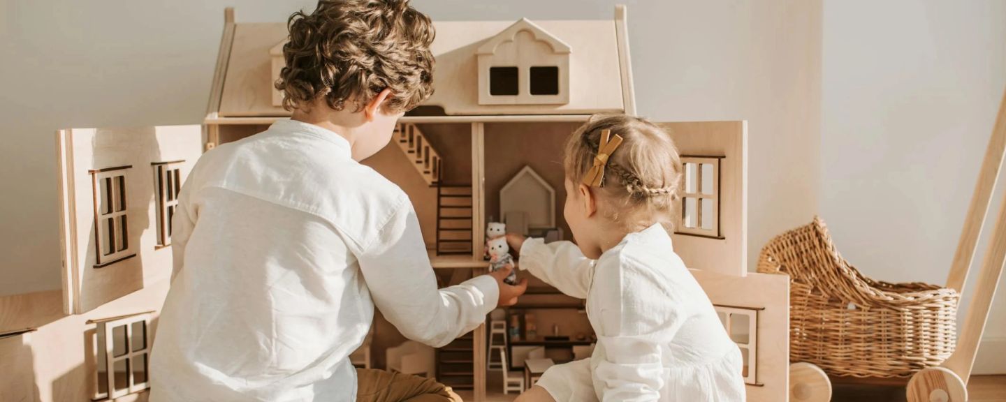 Two children in white clothing playing with a wooden dollhouse