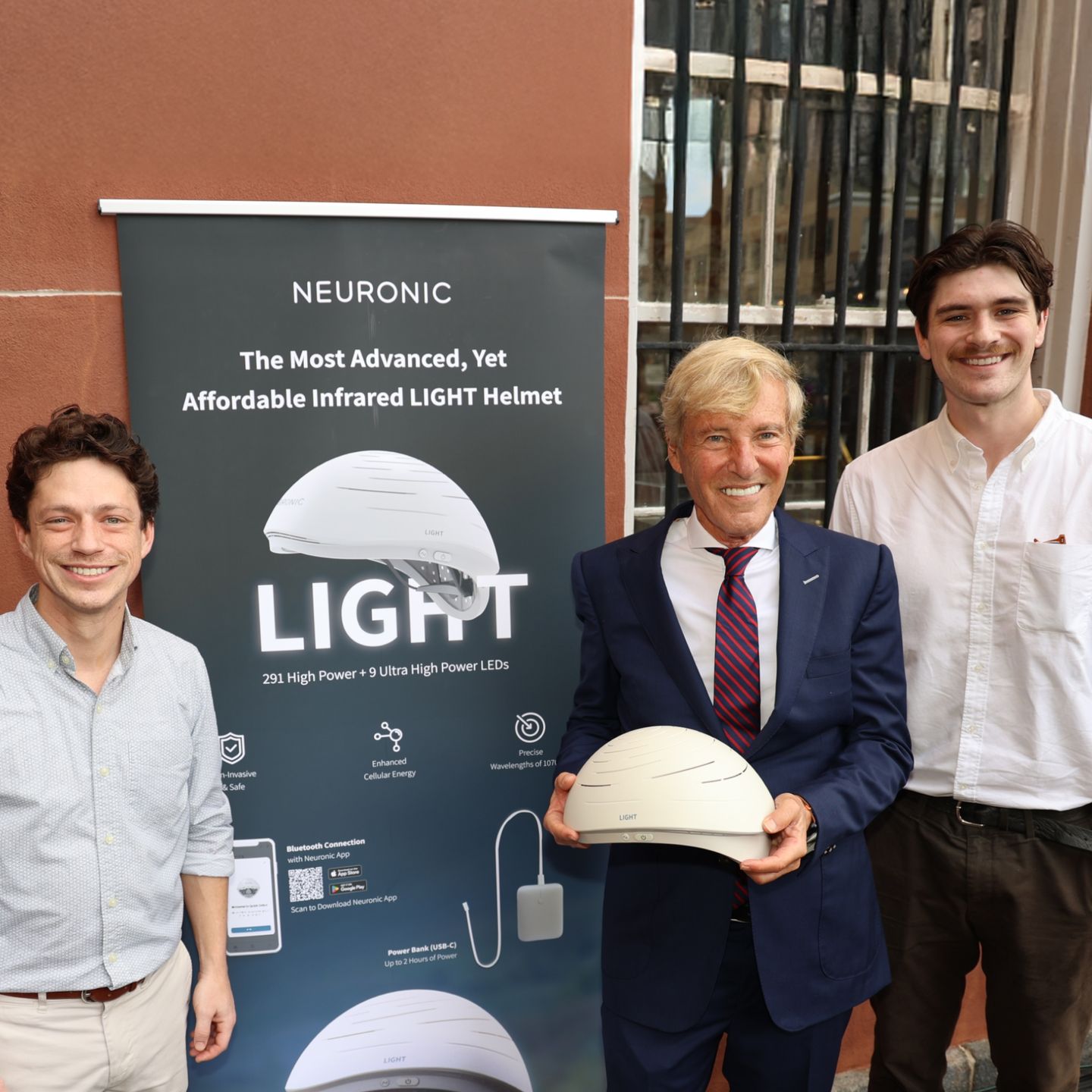Three men standing next to a Neuronic LIGHT product banner, with the center man holding a white Neuronic Helmet