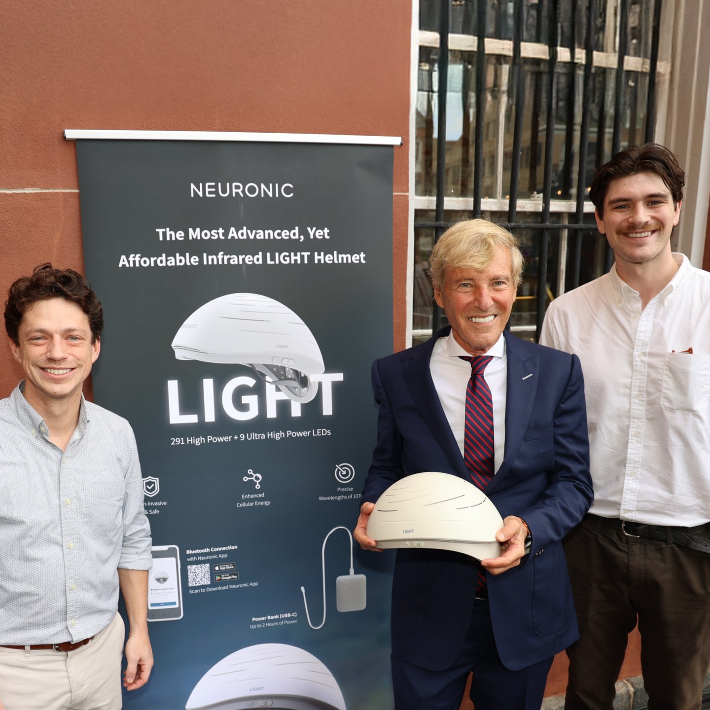 Three men standing next to a Neuronic LIGHT product banner, with the center man holding a white Neuronic Helmet