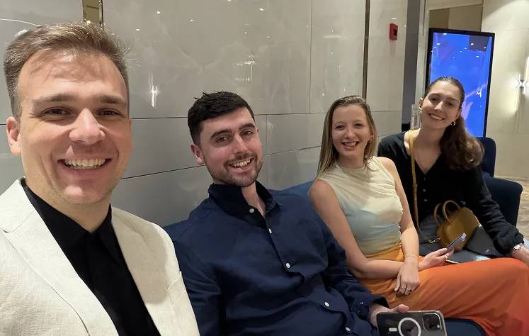 Four people smiling at the camera, seated indoors in front of a glass wall with a blue digital display