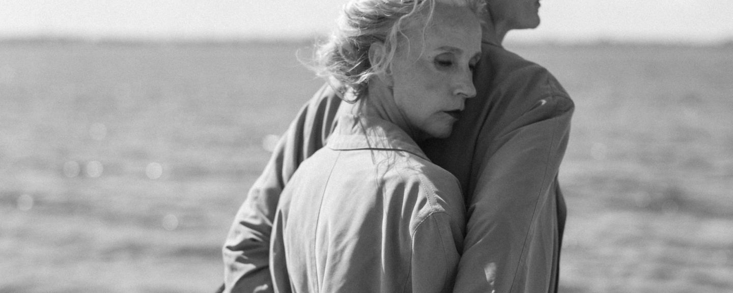 Two people standing together by the ocean in black and white, looking toward the horizon