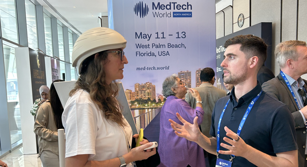 Two people conversing at a MedTech World conference in West Palm Beach, with attendees and banners visible in the background