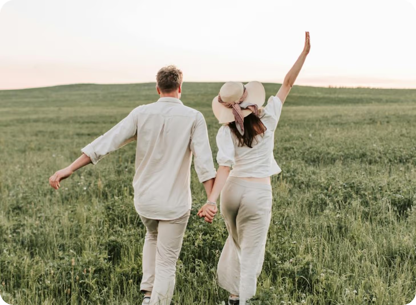 Couple holding hands running through a green field, woman wearing a wide-brimmed hat with arm raised, man in beige shirt viewed from behind