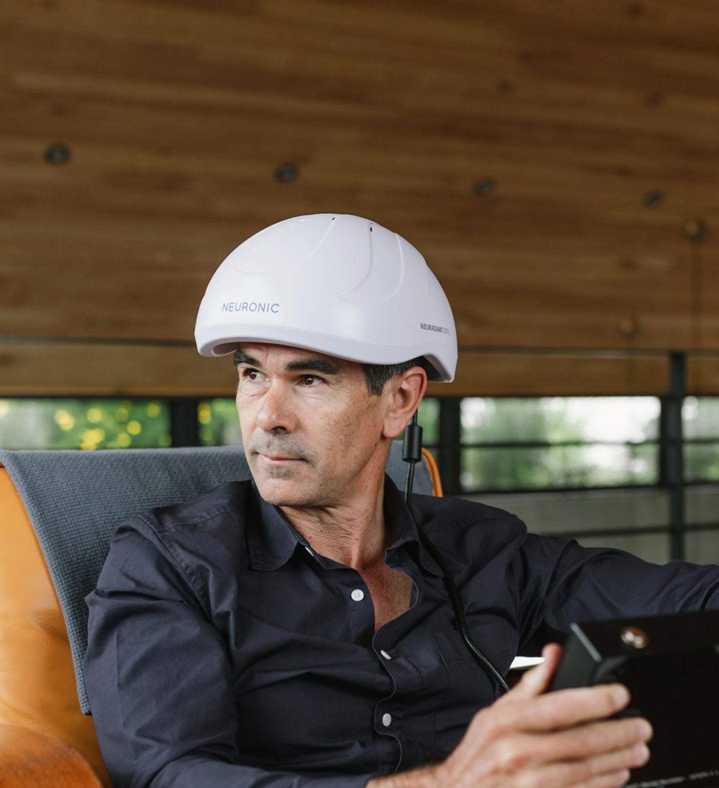 Man wearing Neuronic Helmet sitting in chair, holding mobile device, looking to the side