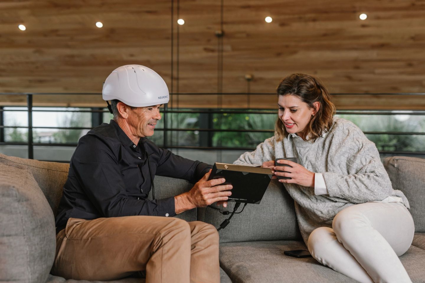 A man wearing the Neuronic LIGHT helmet showing a tablet to a woman sitting beside him on a sofa.