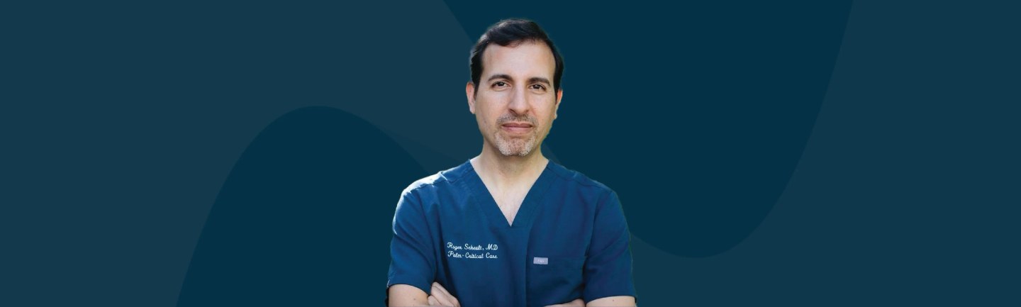 Man in dark blue medical scrubs with embroidered credentials, arms crossed, against teal background
