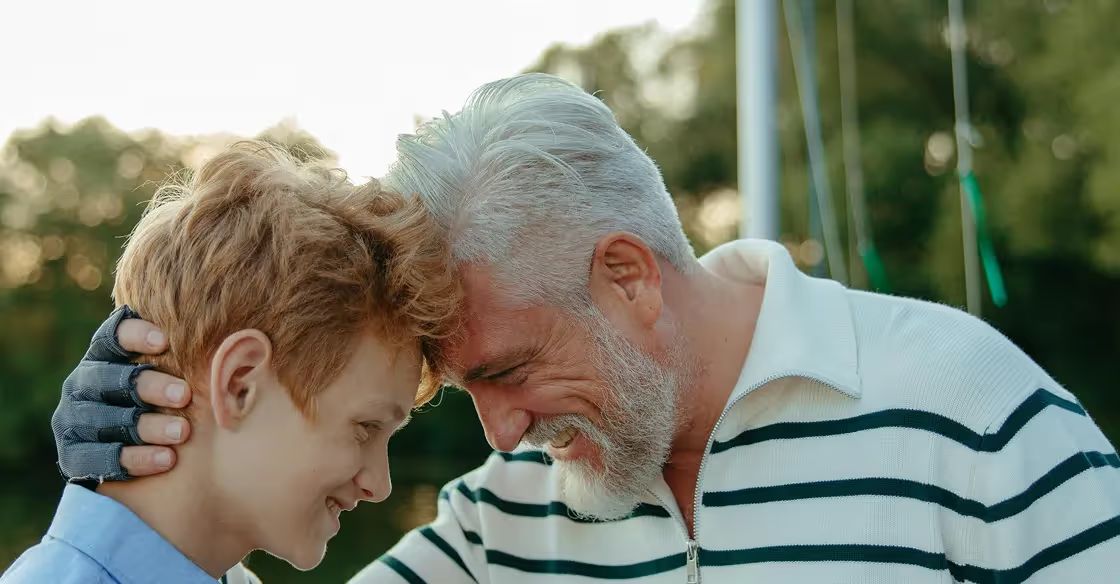 Older man and young boy facing each other outdoors, man's hand on boy's shoulder, both smiling