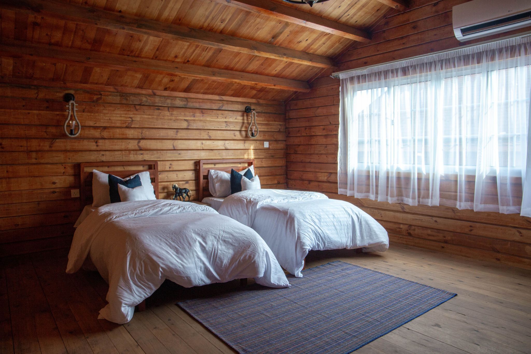 A rustic log cabin bedroom featuring two twin beds with white duvets, wooden walls, a beamed ceiling, and a large window with sheer curtains.