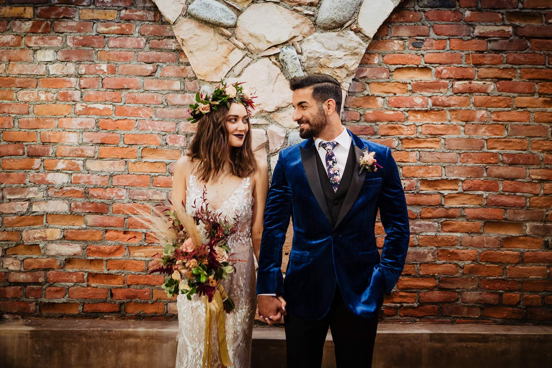 Bride in a flower crown and groom in a blue velvet tuxedo holding hands in front of a brick and stone wall