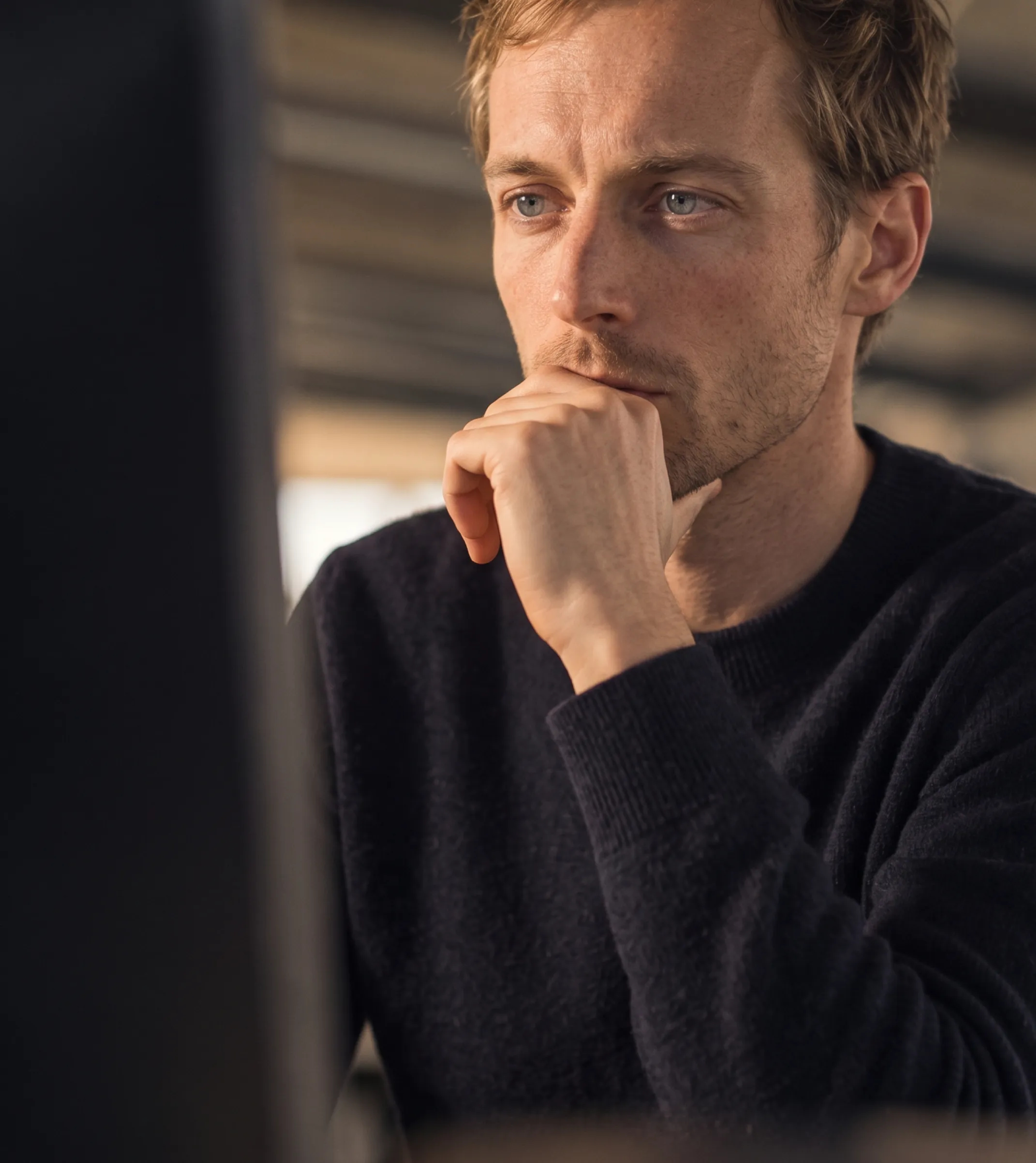 A man in a dark sweater looking intently at a screen, chin resting on his hand