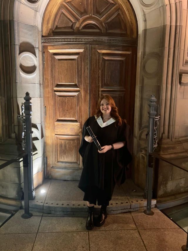 Graduate (Kim O'Neill) in academic dress holding a diploma tube and smiling outside the graduation venue.