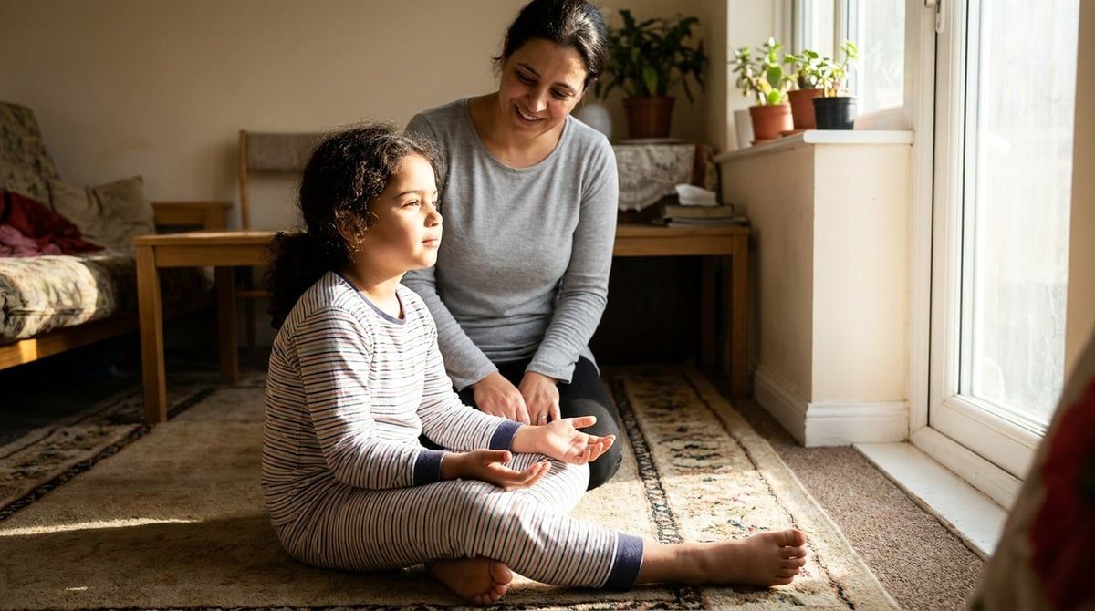 Niña en pijama sentada en una alfombra junto a una ventana con las cortinas abiertas, con la luz de la mañana en la cara, y su madre arrodillada detrás, fotografía natural