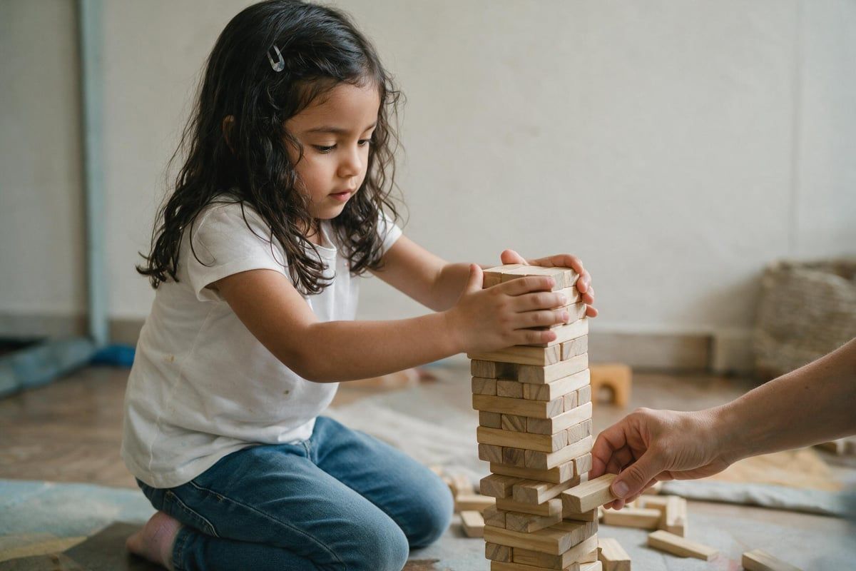 Bambina di circa cinque anni che costruisce con i mattoncini mentre una mano adulta tiene fermo un pezzo della base senza posizionarlo