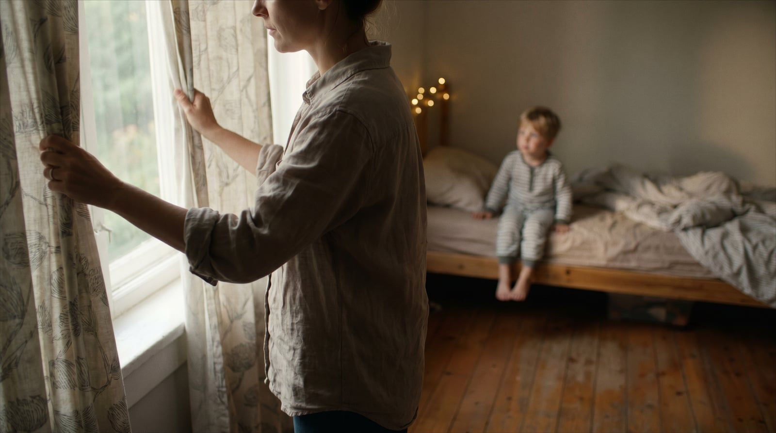 Madre cerrando las cortinas para atenuar la luz del atardecer de verano mientras su hijo pequeño se prepara para dormir, fotografía natural