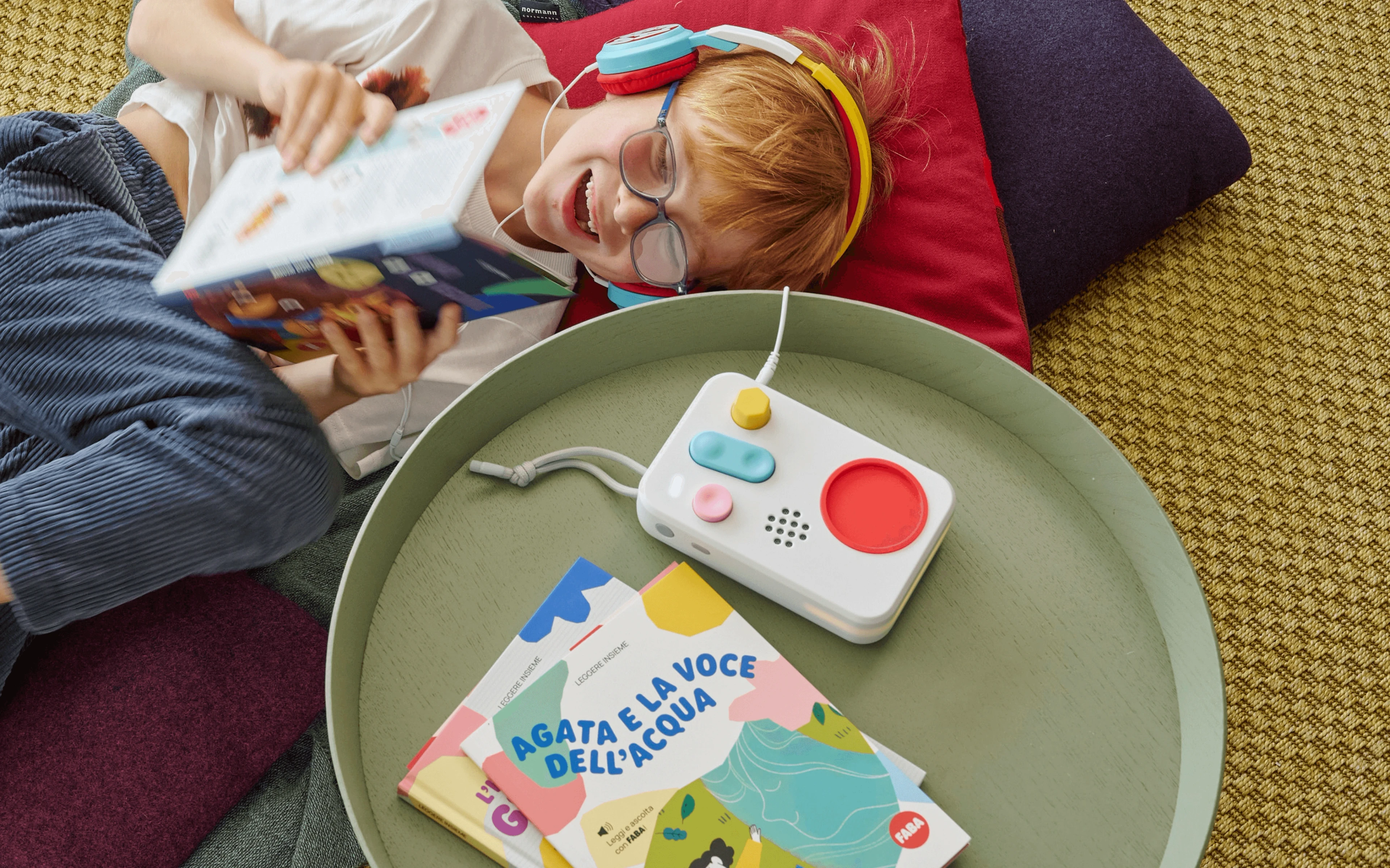 Child lying on cushions with headphones on, listening to the FABA+ speaker while reading a FABA book