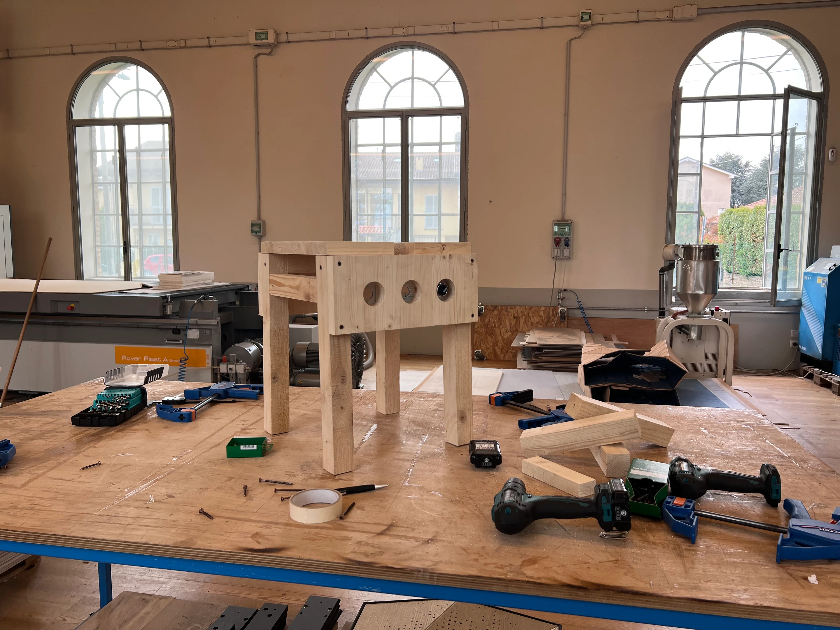A timber frame taking shape on a workshop bench under arched windows