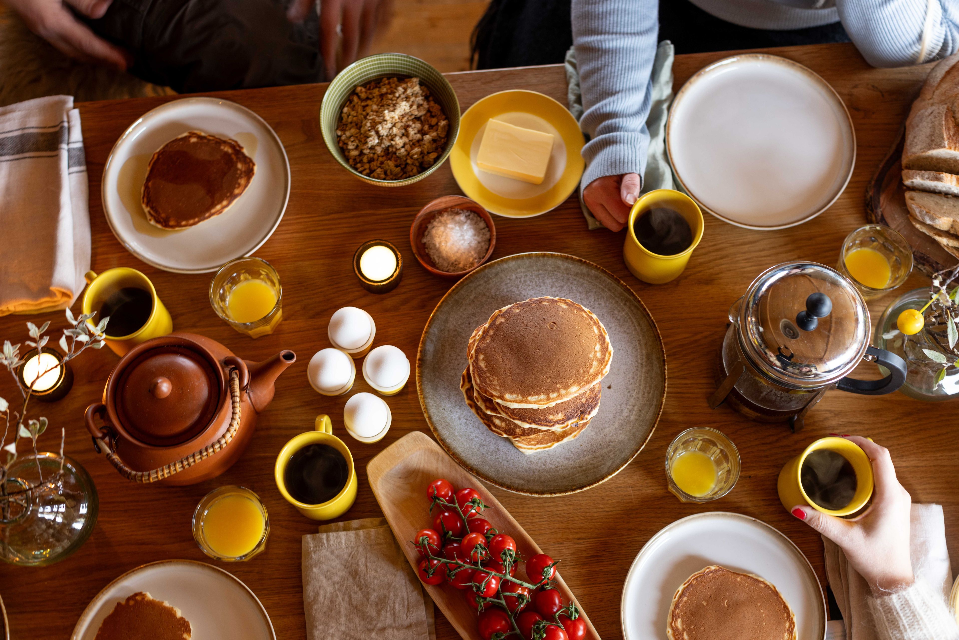 Pannekaker, kaffekopper, tomater og kokte egg på et frokostbord.